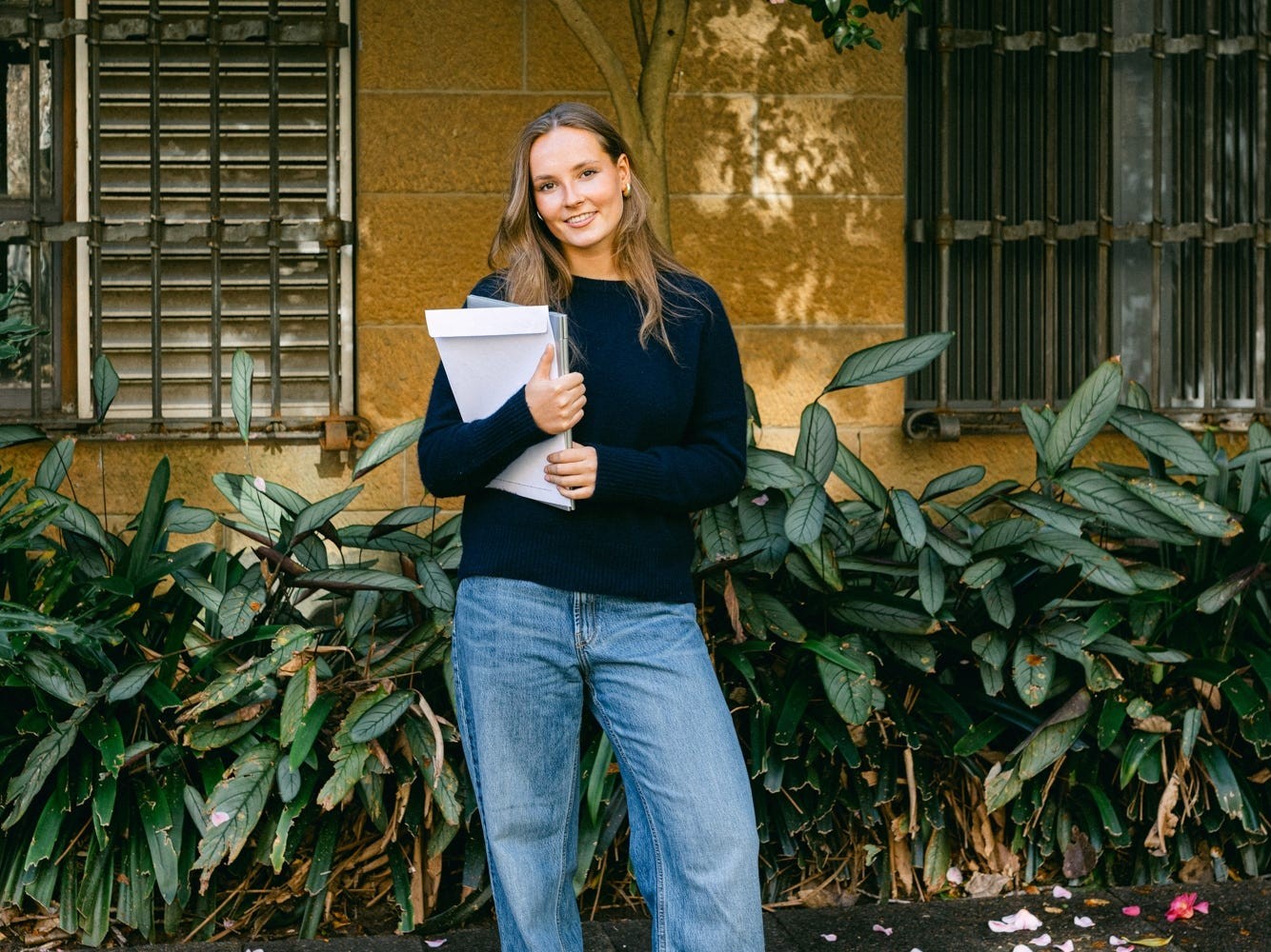Young woman holding documents, smiling outdoors in front of a flowering tree and greenery.
