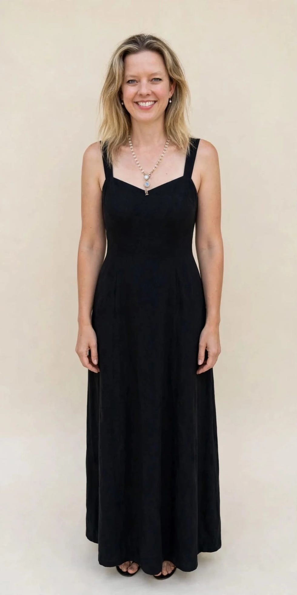Woman smiling in a black dress with a necklace, standing against a neutral background.