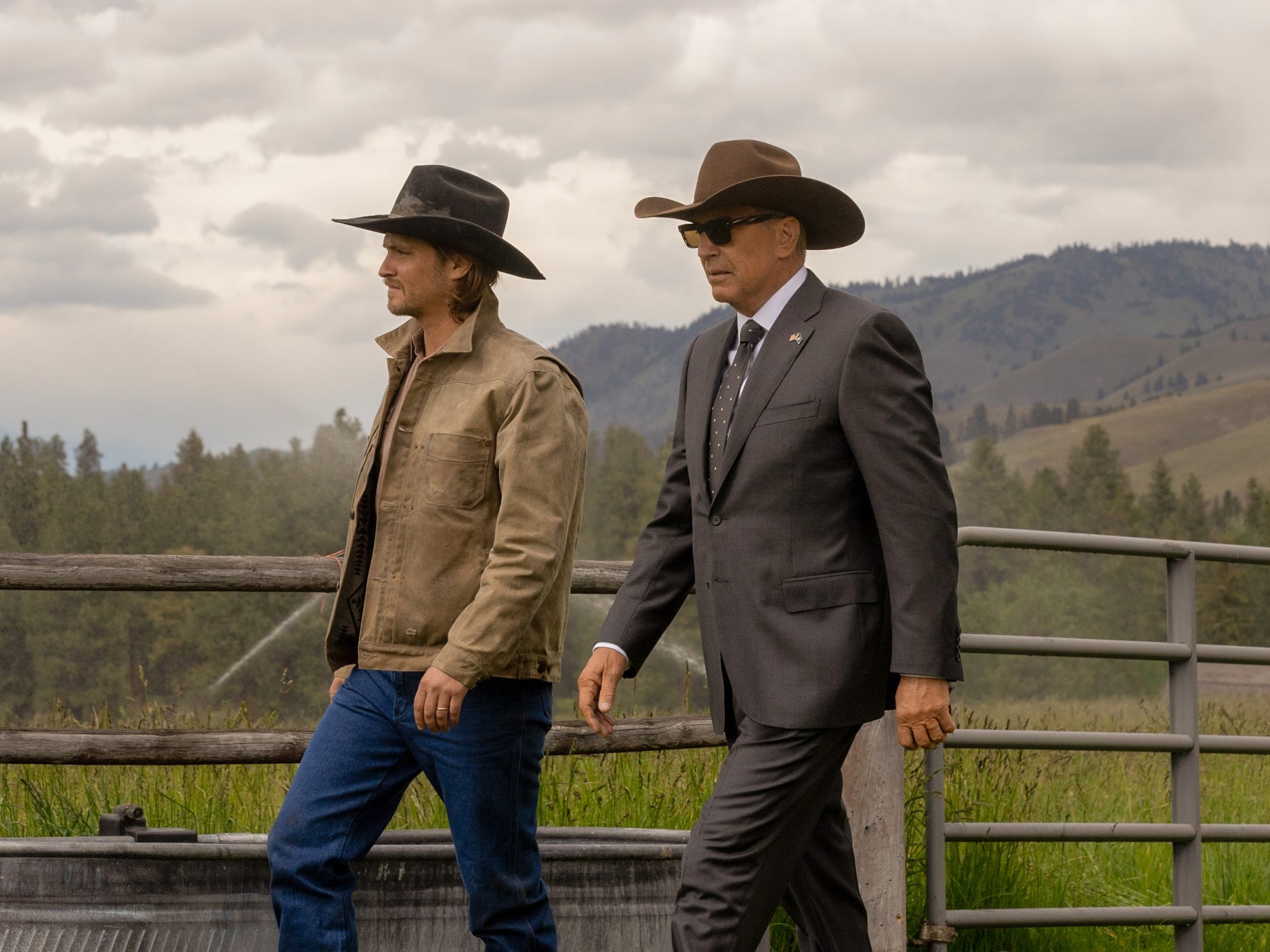Two men in cowboy hats walking along a ranch fence against a cloudy sky and green landscape.