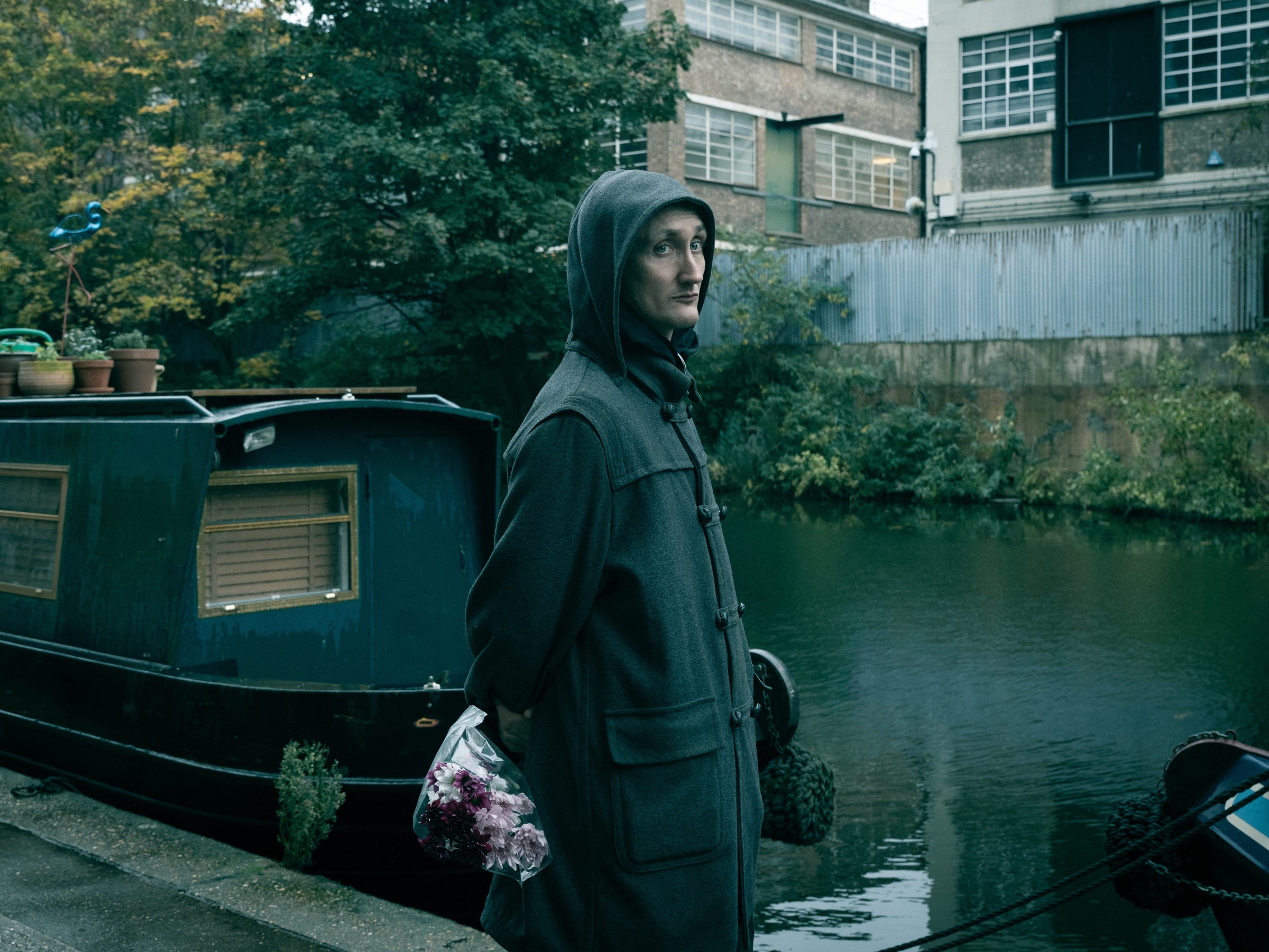 A person with a hooded coat holds flowers while standing by a canal near a green narrowboat and urban buildings.
