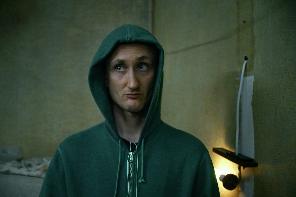 Man in a green hoodie looking up with a neutral expression in a dimly lit room.