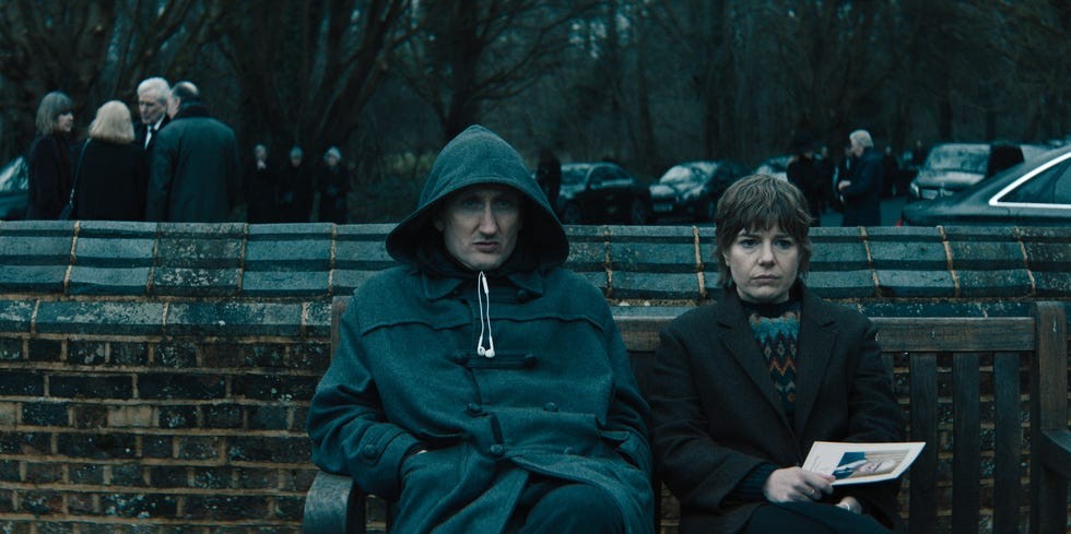Two individuals sitting on a bench with a somber expression in a gloomy outdoor setting with a gathering in the background.