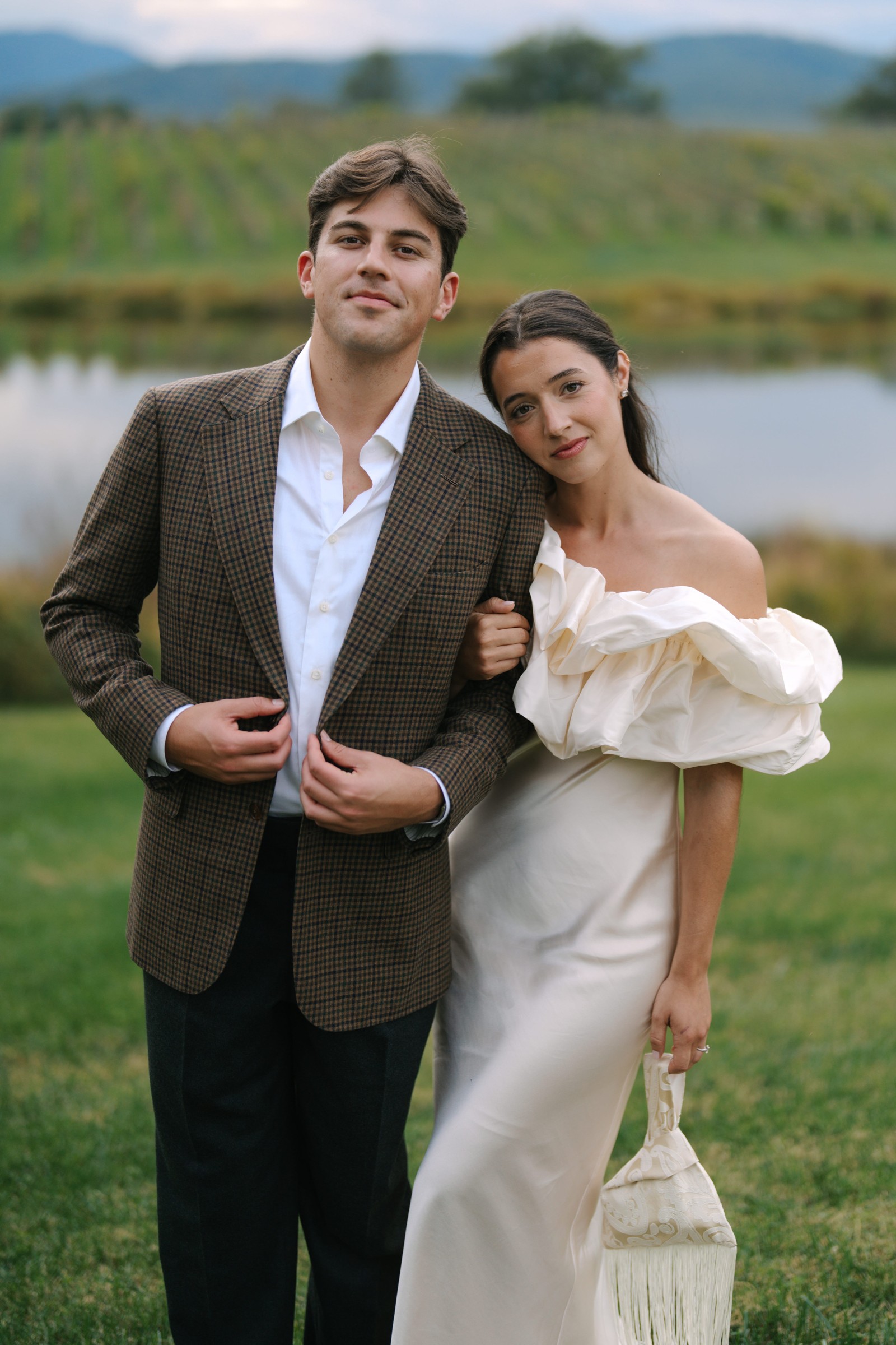 A couple poses near a pond, the man in a checkered blazer and the woman in an elegant off-the-shoulder dress.