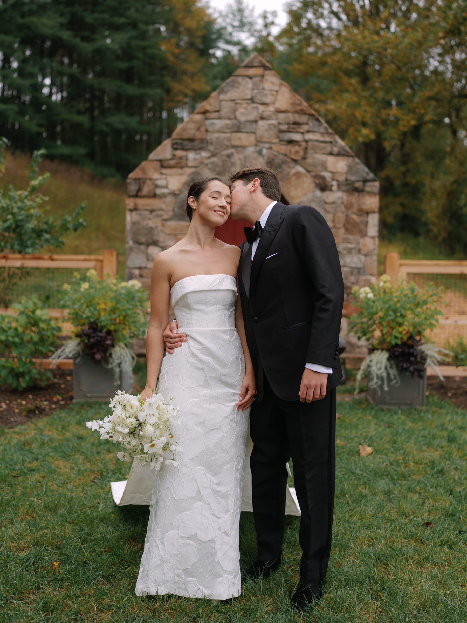 Bride and groom share a tender moment in a garden setting, with the bride holding a bouquet of white flowers.