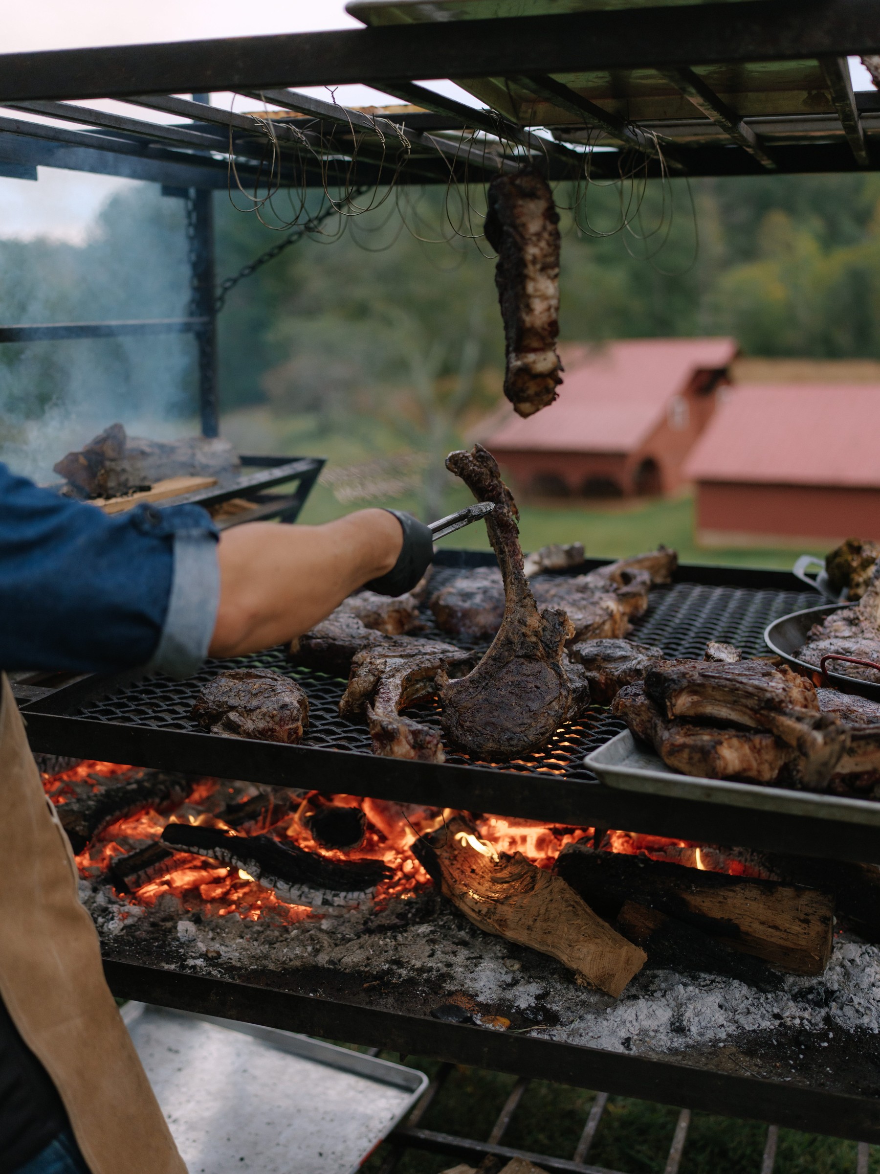 Grilling various cuts of meat over an open flame with smoke rising in a rustic outdoor setting.