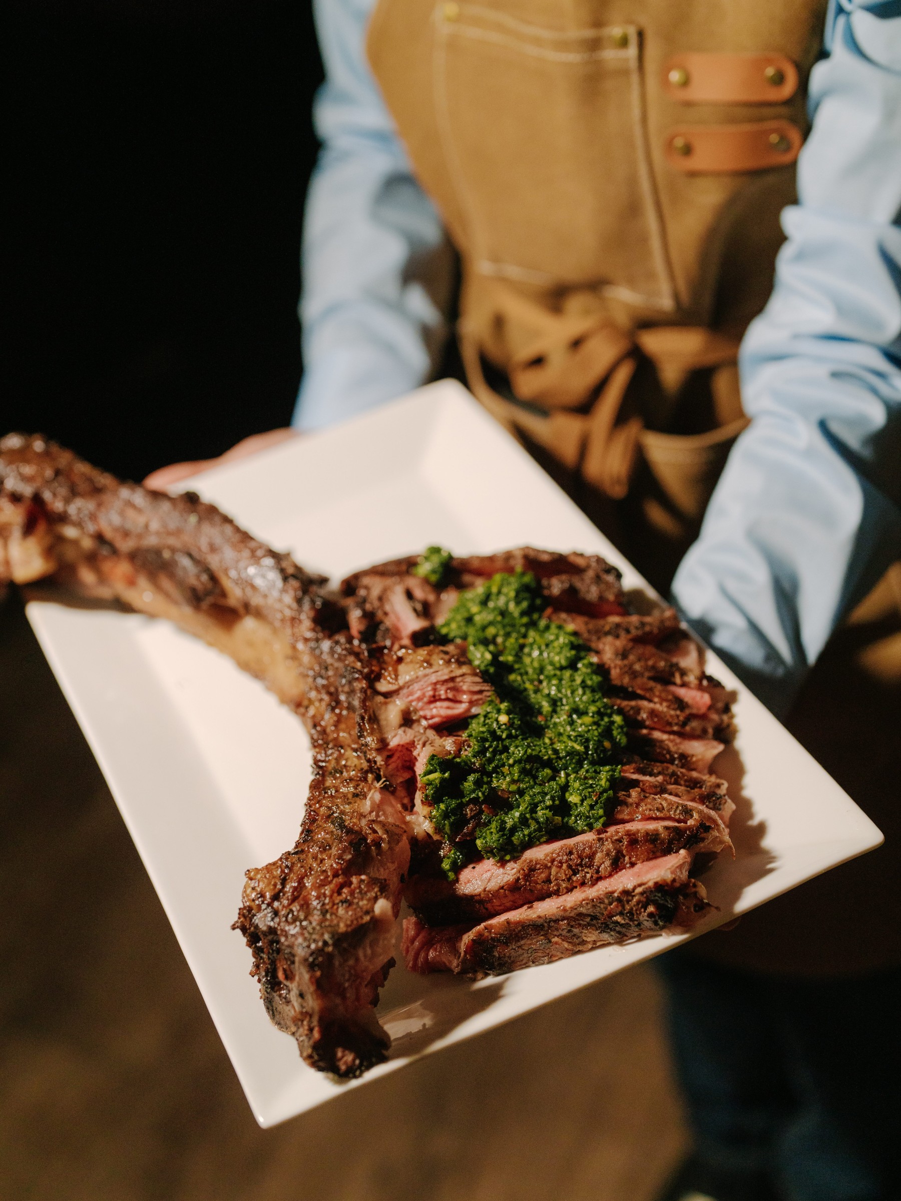 A platter of sliced steak topped with green chimichurri sauce, presented by a person in an apron.