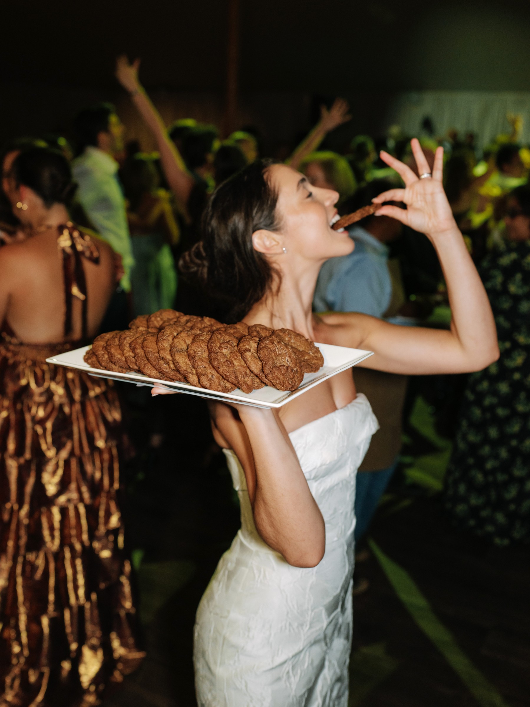 Woman in a white dress joyfully holding a tray of cookies while enjoying a lively party atmosphere.