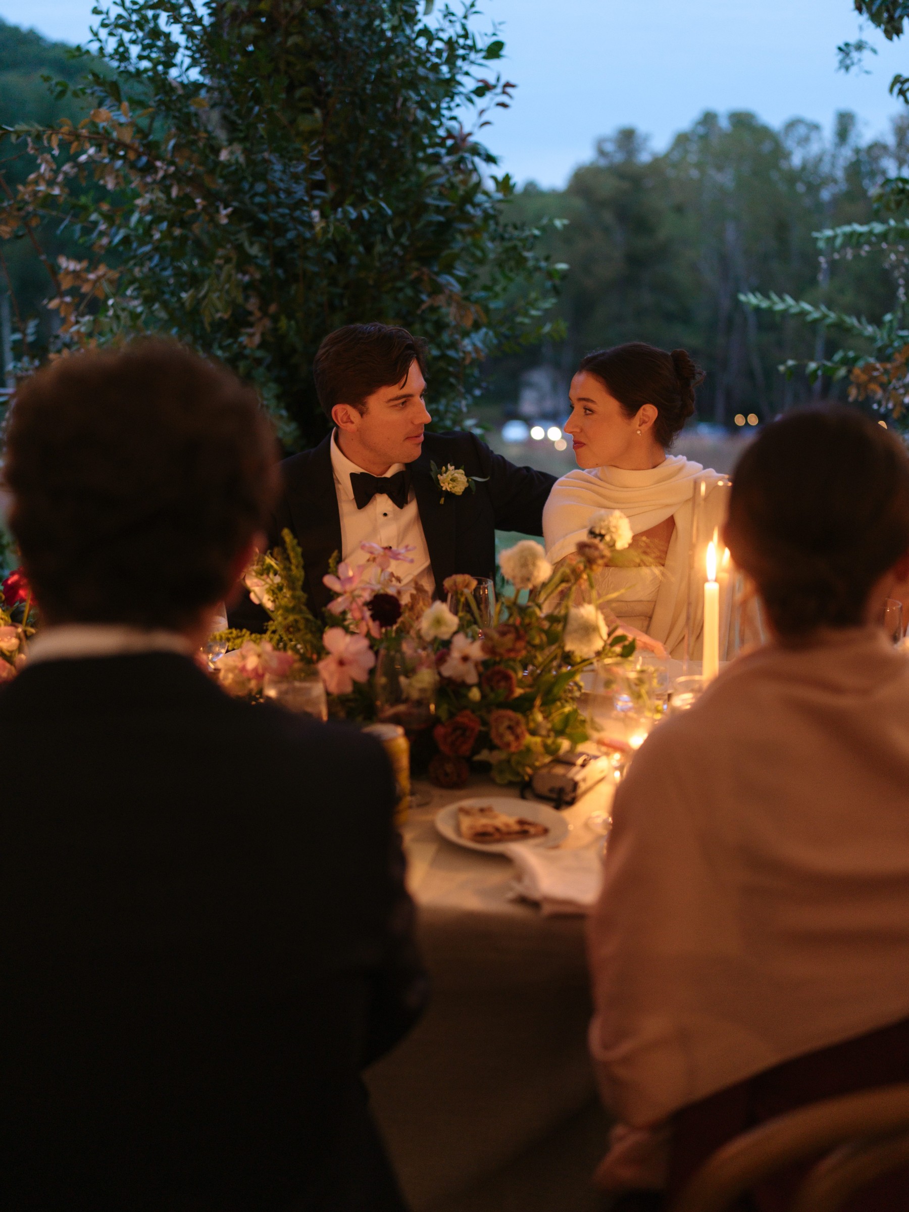 A couple enjoying an intimate dinner surrounded by candles and flowers, with guests seated in the foreground.