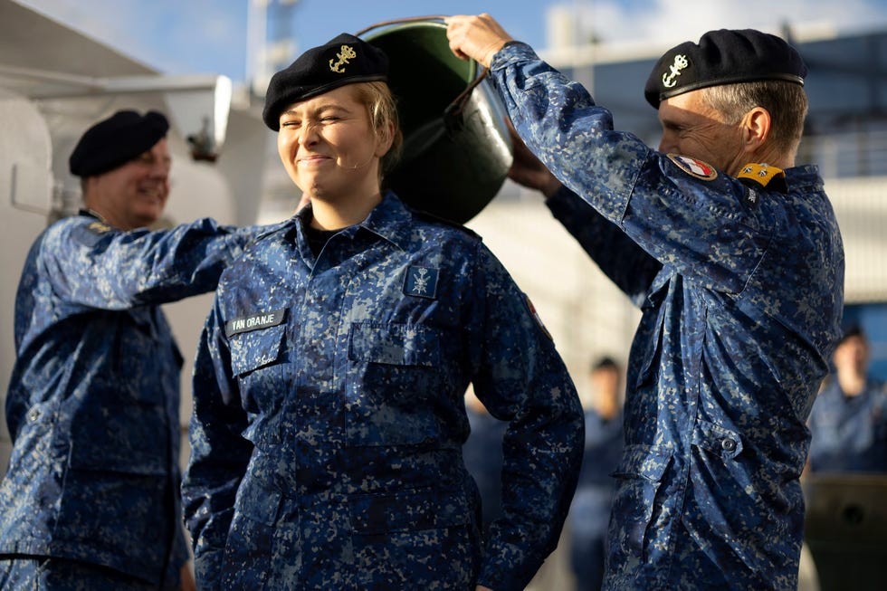 A person in military uniform smiles while having a helmet placed on their head, with others in the background.