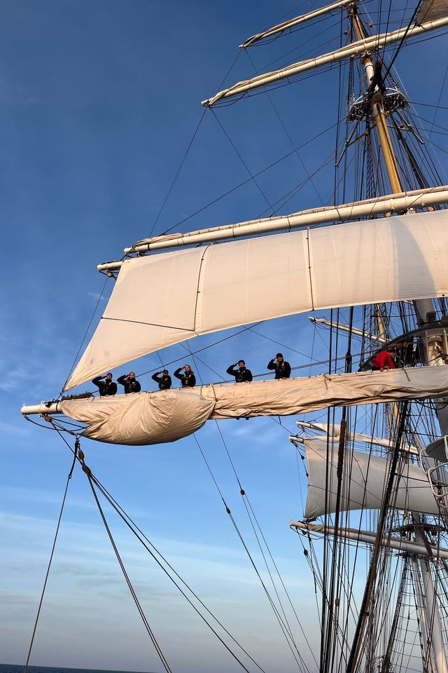 Crew members saluting from the sails of a tall ship against a blue sky.
