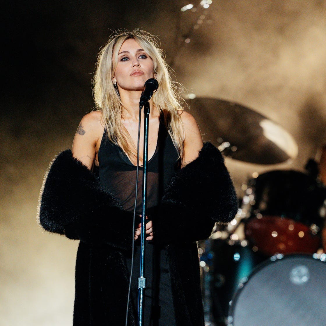 A singer performing on stage, microphone in hand, with a drummer visible in the background and fog filling the scene.