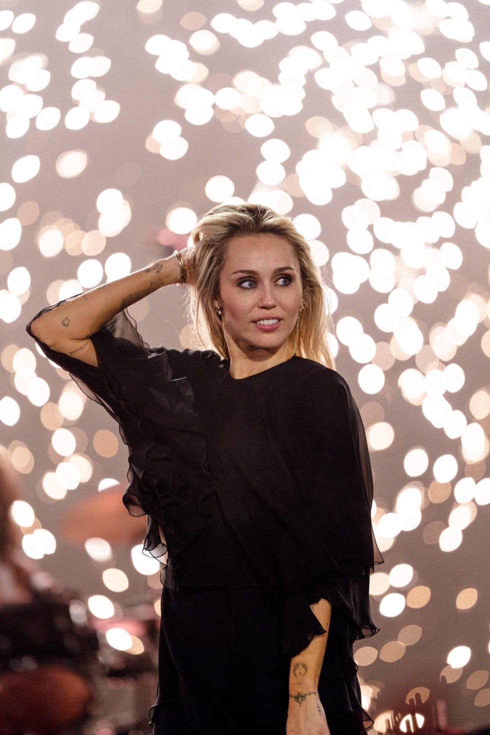 A woman in a black dress poses confidently against a backdrop of bright, blurred lights.