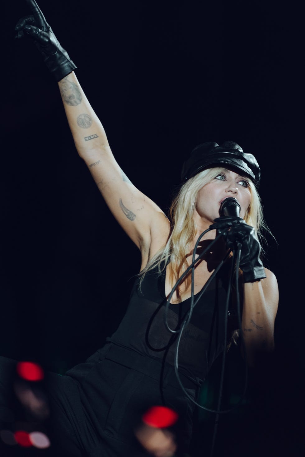 A singer performs energetically on stage, holding a microphone and gesturing with a raised arm, wearing a black outfit and gloves.