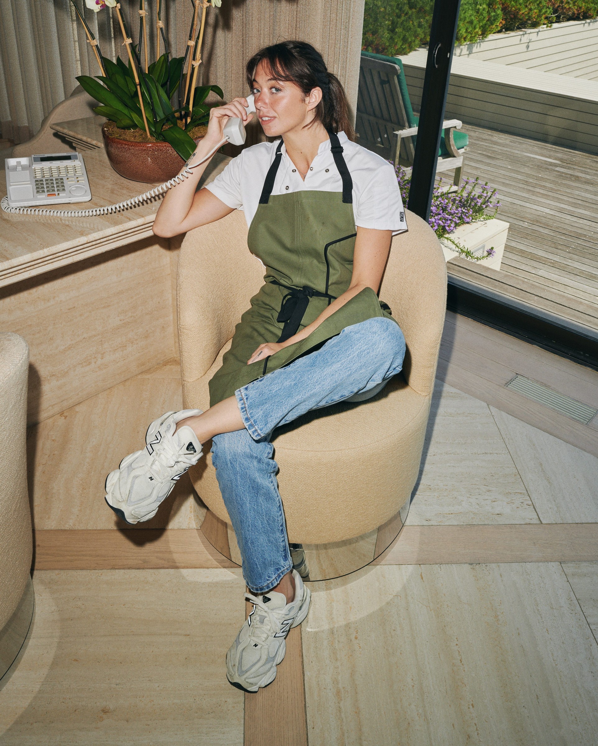 A woman in an apron sits on a chair, talking on a phone near a pot of orchids with a sunny outdoor view.