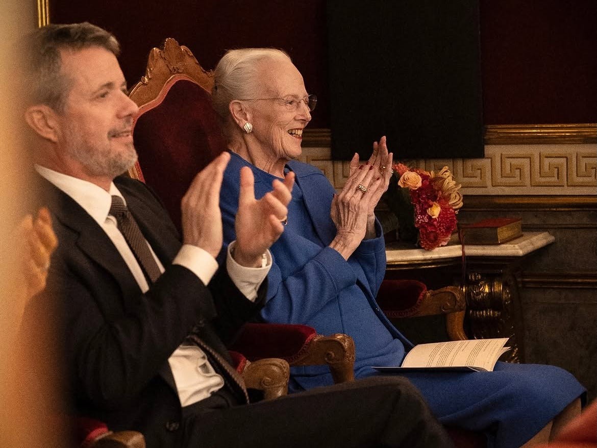 Two individuals, one in a suit and one in a blue outfit, sitting and clapping in a formal setting.