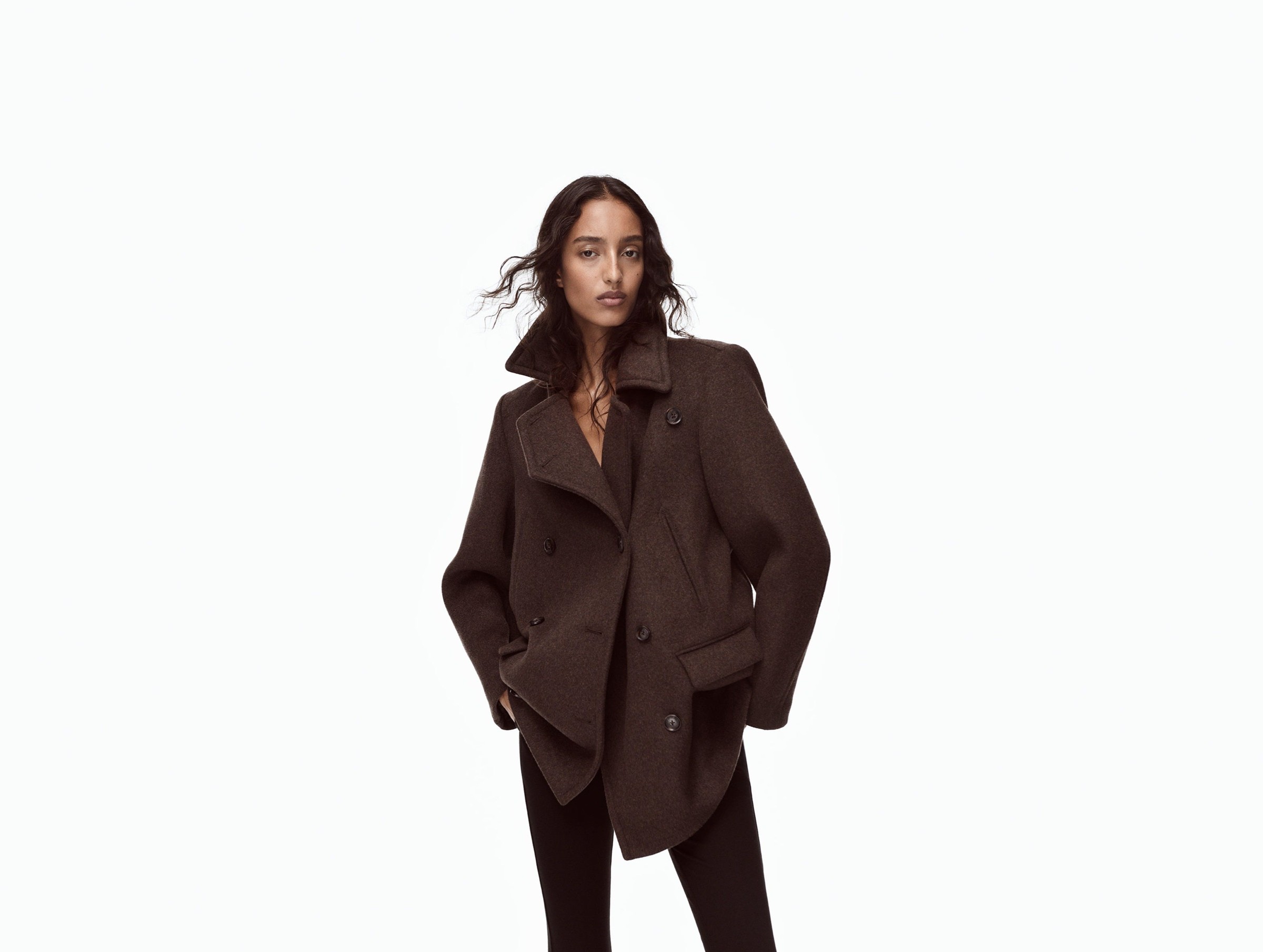 Model wearing a dark brown oversized coat with a high collar, posing confidently against a plain background.