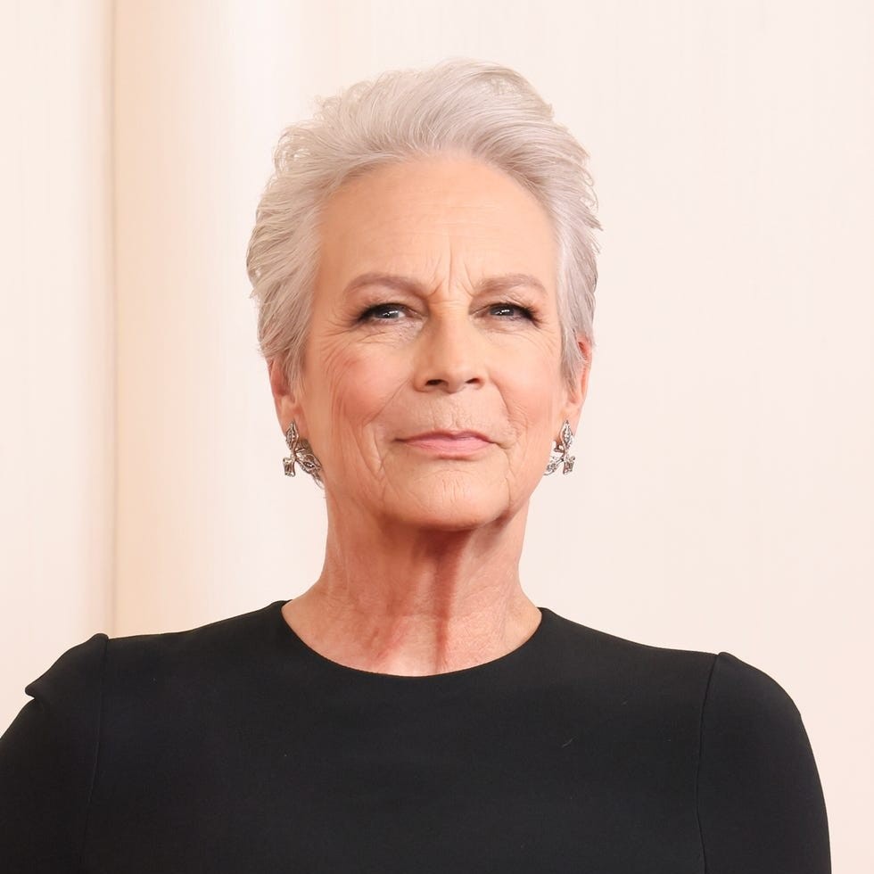 Woman with short gray hair wearing a black dress and silver earrings, confidently posing against a light background.