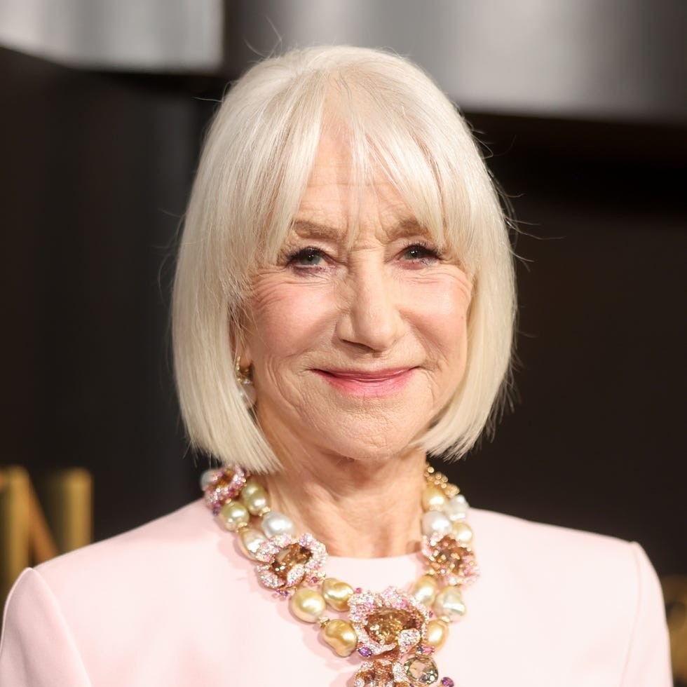 Older woman with short white hair wearing a pink dress and a statement necklace with pearls and gemstones.