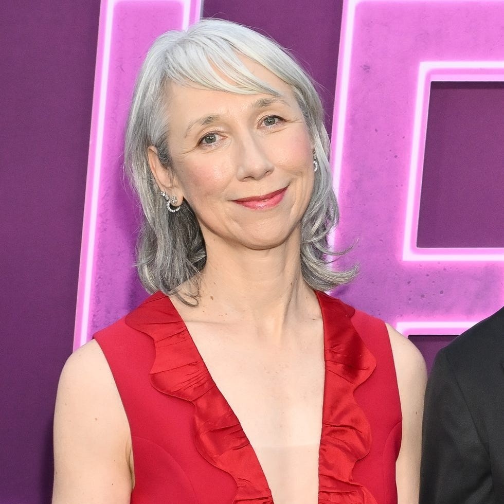 Woman with short gray hair wearing a red dress with ruffles, smiling against a purple backdrop at an event.