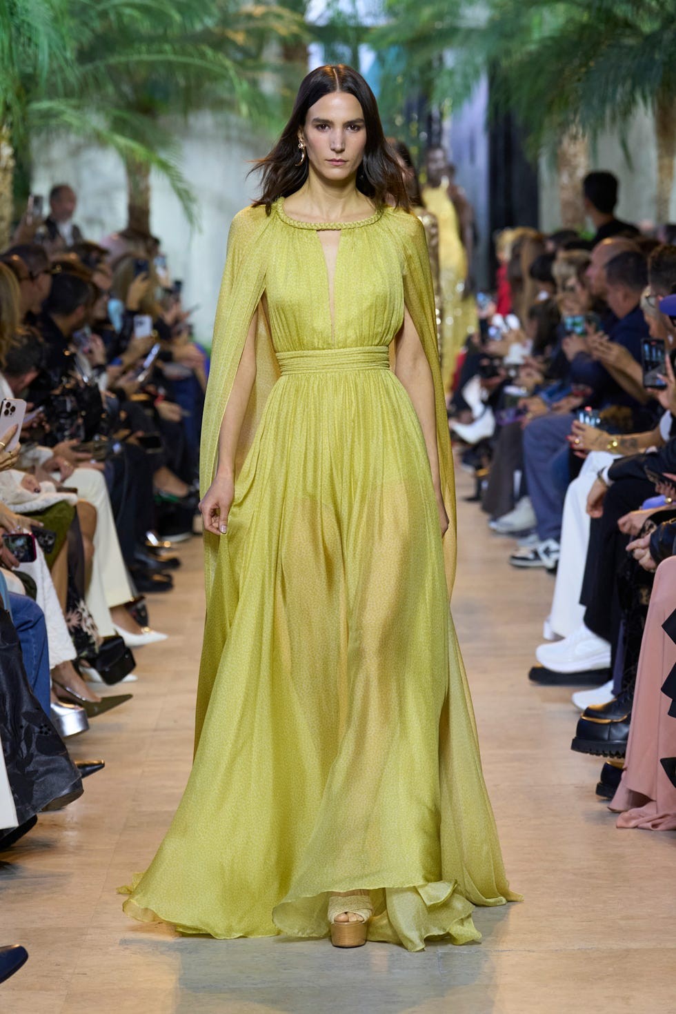 Model walking the runway in a flowing yellow gown with a cape, surrounded by an audience and green plants.