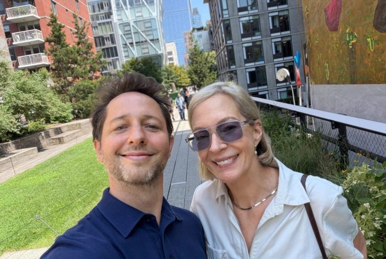 Two smiling individuals taking a selfie in an urban park with modern buildings in the background.