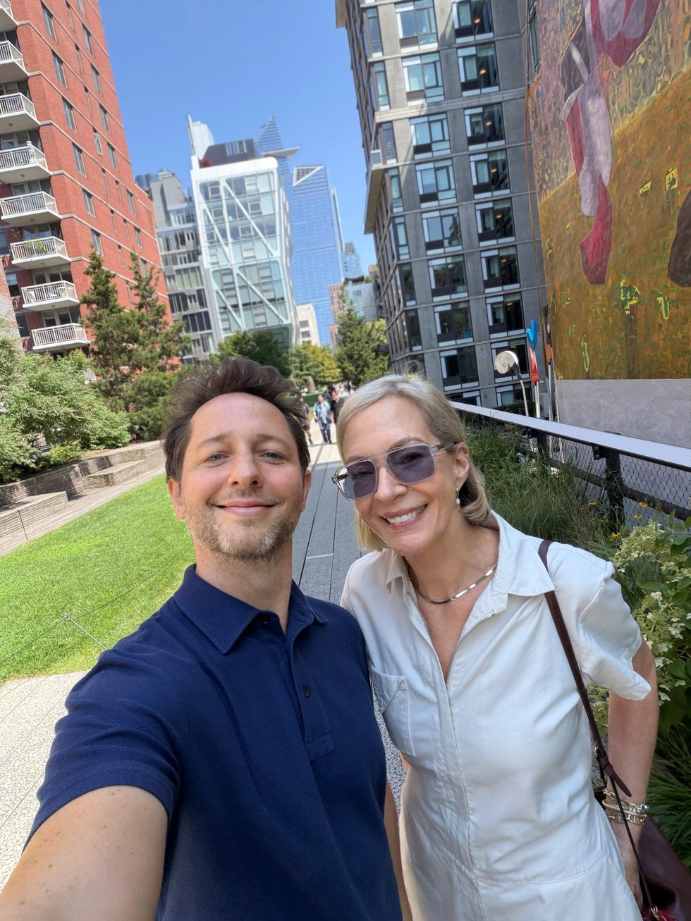 Two smiling individuals taking a selfie in an urban park with modern buildings in the background.