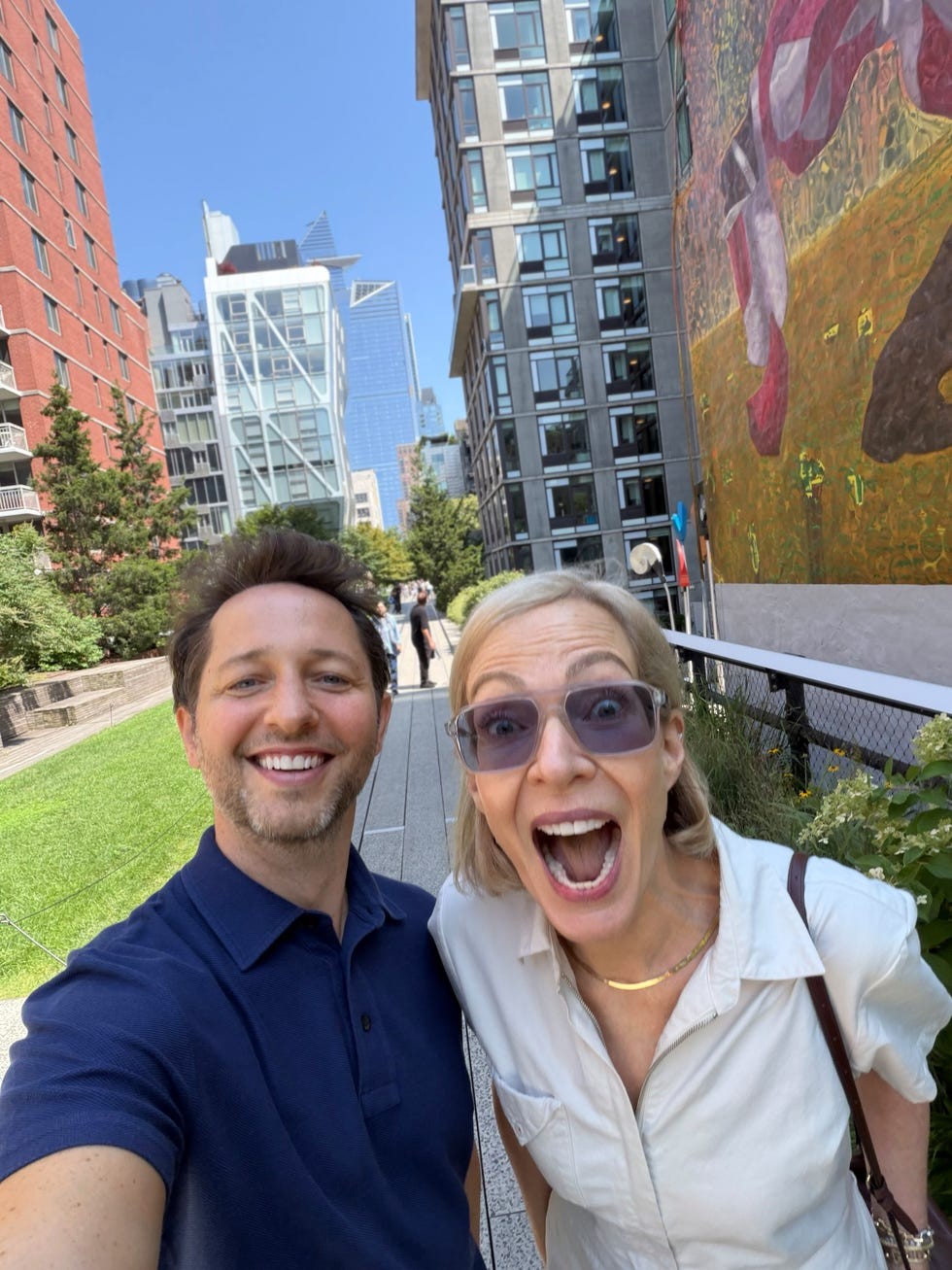 Two smiling individuals posing for a selfie outdoors, with buildings and greenery in the background.