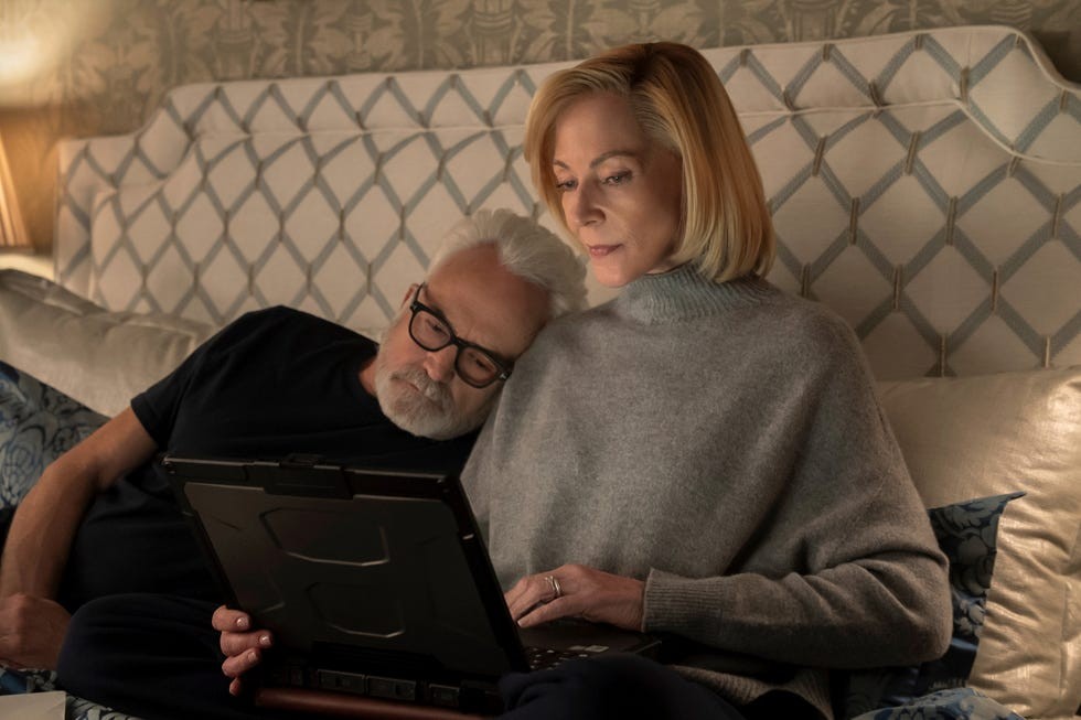 A woman and man sit together on a bed, looking intently at a laptop in a cozy, softly lit bedroom.