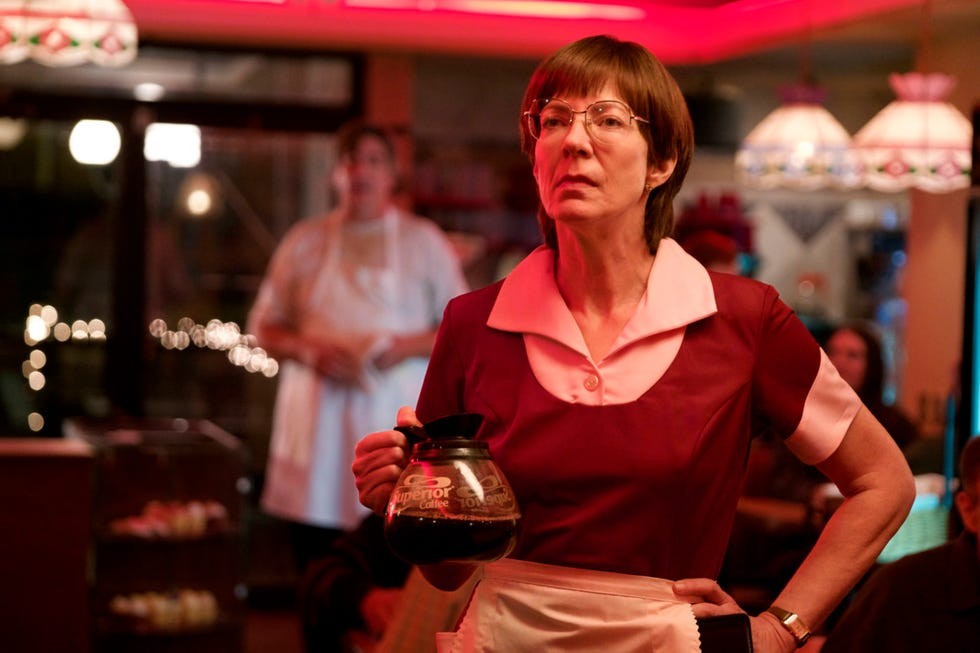 A waitress in a diner holding a coffee pot, with a blurred figure in the background.
