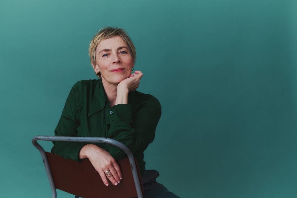 A woman with short hair in a green blouse resting her chin on her hand, seated against a teal background.