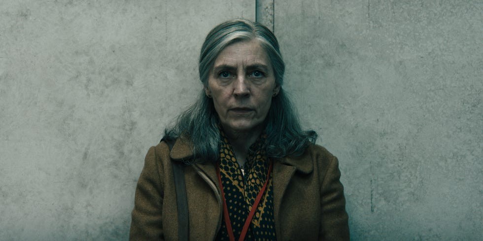 A woman with gray hair and a serious expression sits against a textured gray wall, wearing a brown coat and patterned scarf.