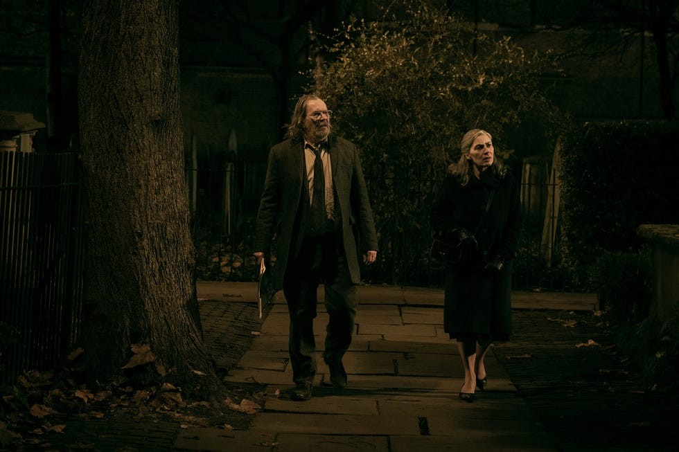 Two individuals walking on a dimly lit street surrounded by trees and foliage at night.