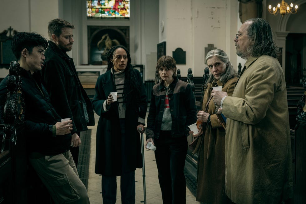 Six people discussing in a church, holding cups, with stained glass windows and wooden pews in the background.