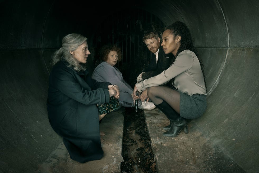 Four people sitting in a dark tunnel discussing something serious, with a muddy ground and a grating in the background.