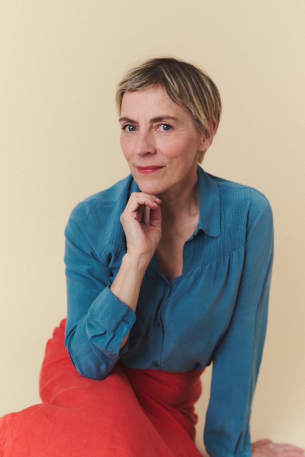 Woman with short hair in a blue shirt and red skirt sitting thoughtfully against a light beige background.