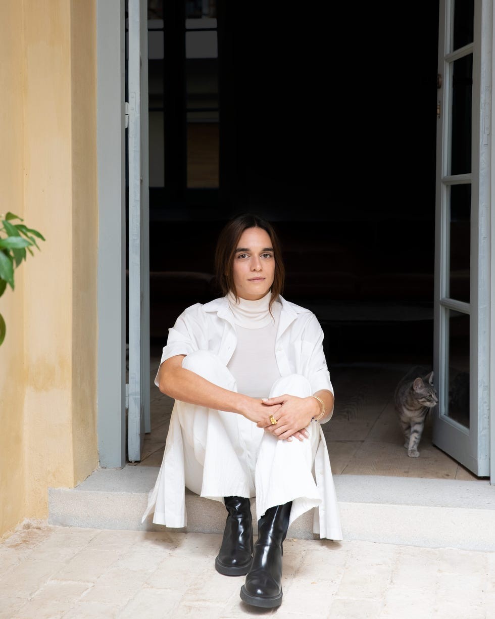 A person in stylish attire sitting on steps with a cat nearby, framed by an open doorway and soft, natural lighting.