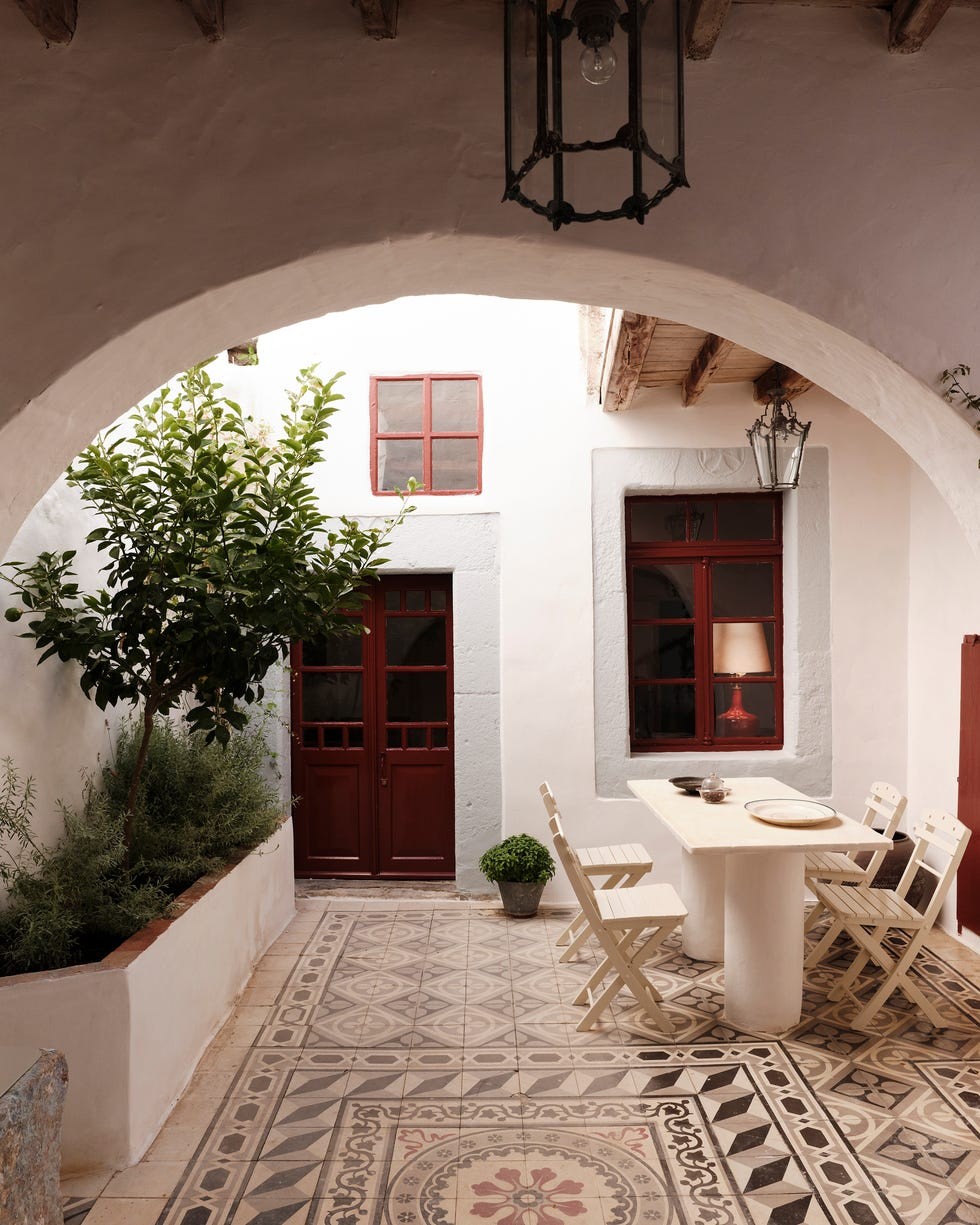A serene courtyard with patterned tiles, a table, chairs, a green plant, and red window frames under soft lighting.