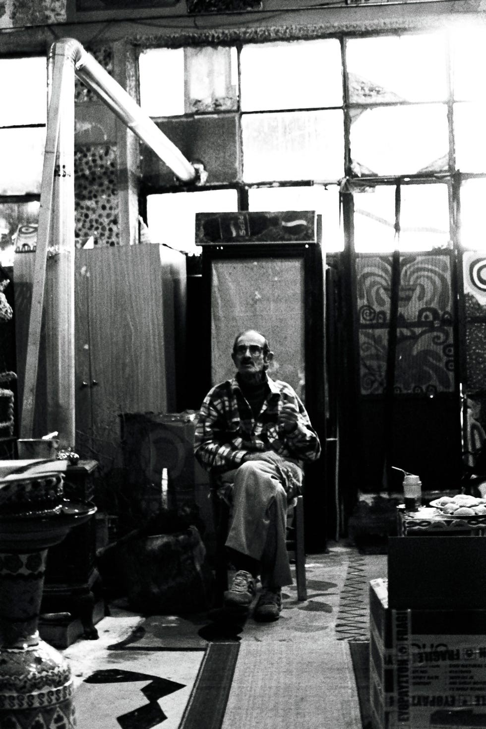 An older man sitting in a dimly lit room surrounded by various objects and artworks.