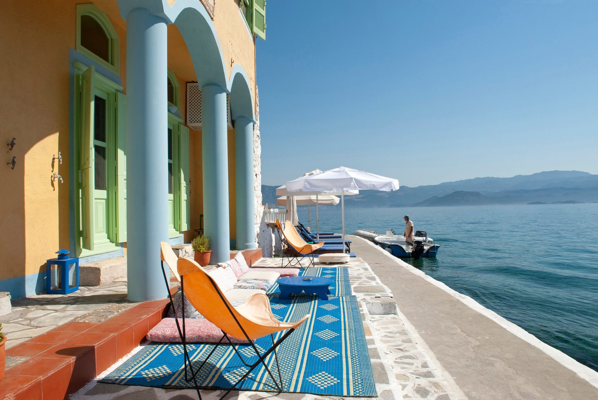 Brightly colored waterfront villa with lounge chairs and a sailboat in a serene landscape.