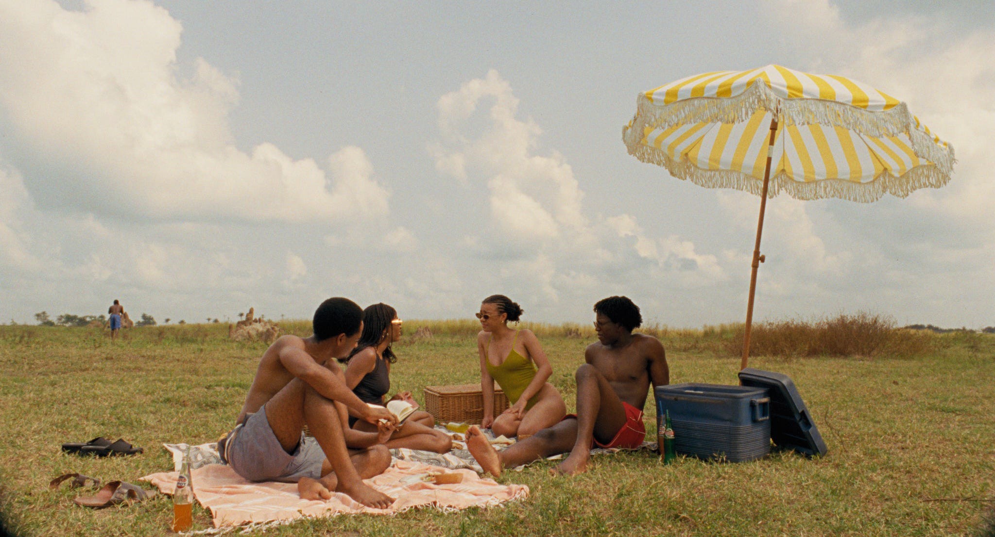 Group of friends enjoying a picnic under a striped umbrella on a grassy field with cloudy skies.
