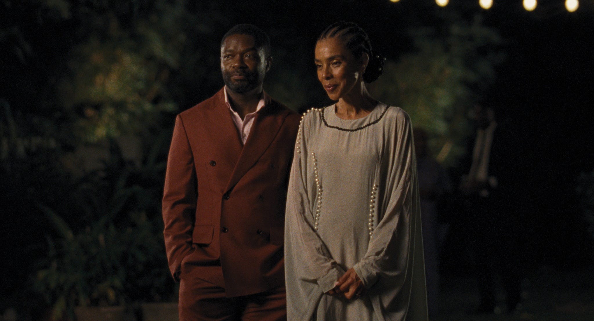 A man in a red suit and a woman in a light dress stand together, smiling in a softly lit outdoor setting.
