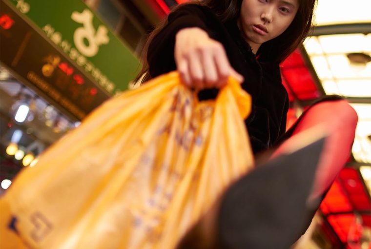 A person in red tights holding an orange bag with a blurred city background illuminated by vibrant lights.