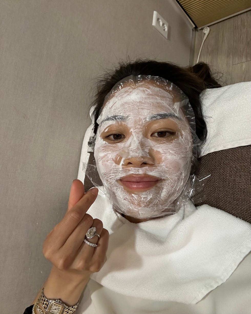 Person with a facial mask and plastic wrap on their face, posing with a heart gesture using one hand.