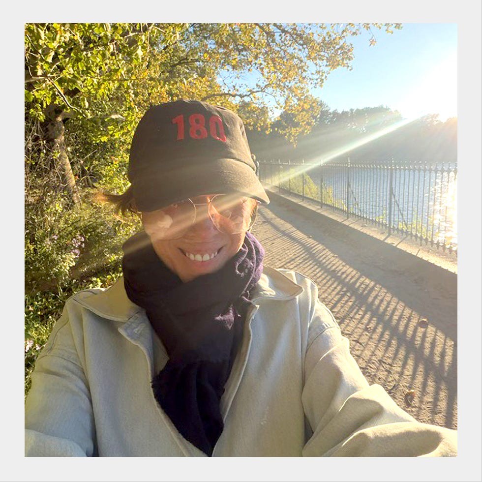 Smiling person wearing a black cap and scarf along a sunlit pathway by a river with trees in the background.