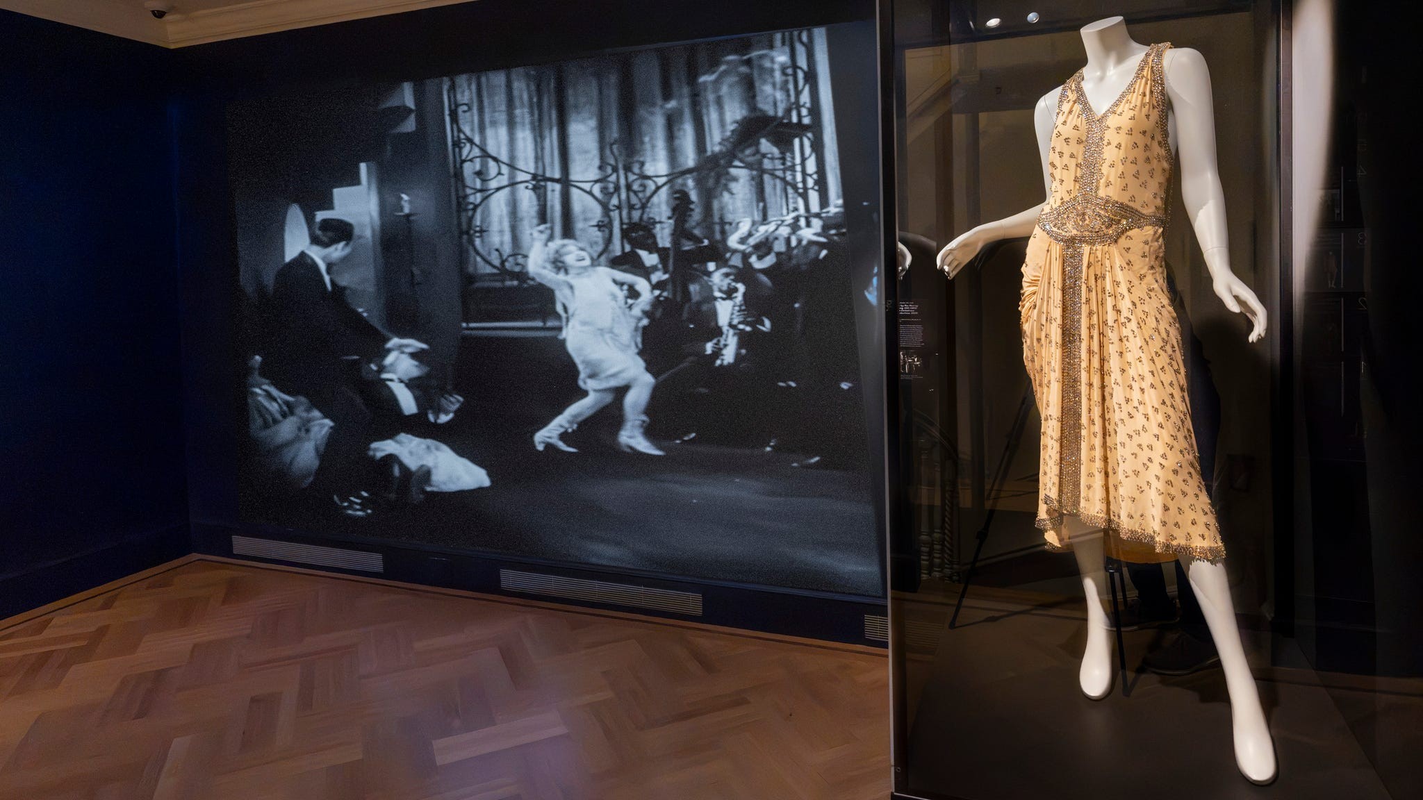 A vintage dress displayed in a glass case with a black and white film backdrop featuring dancing scenes.