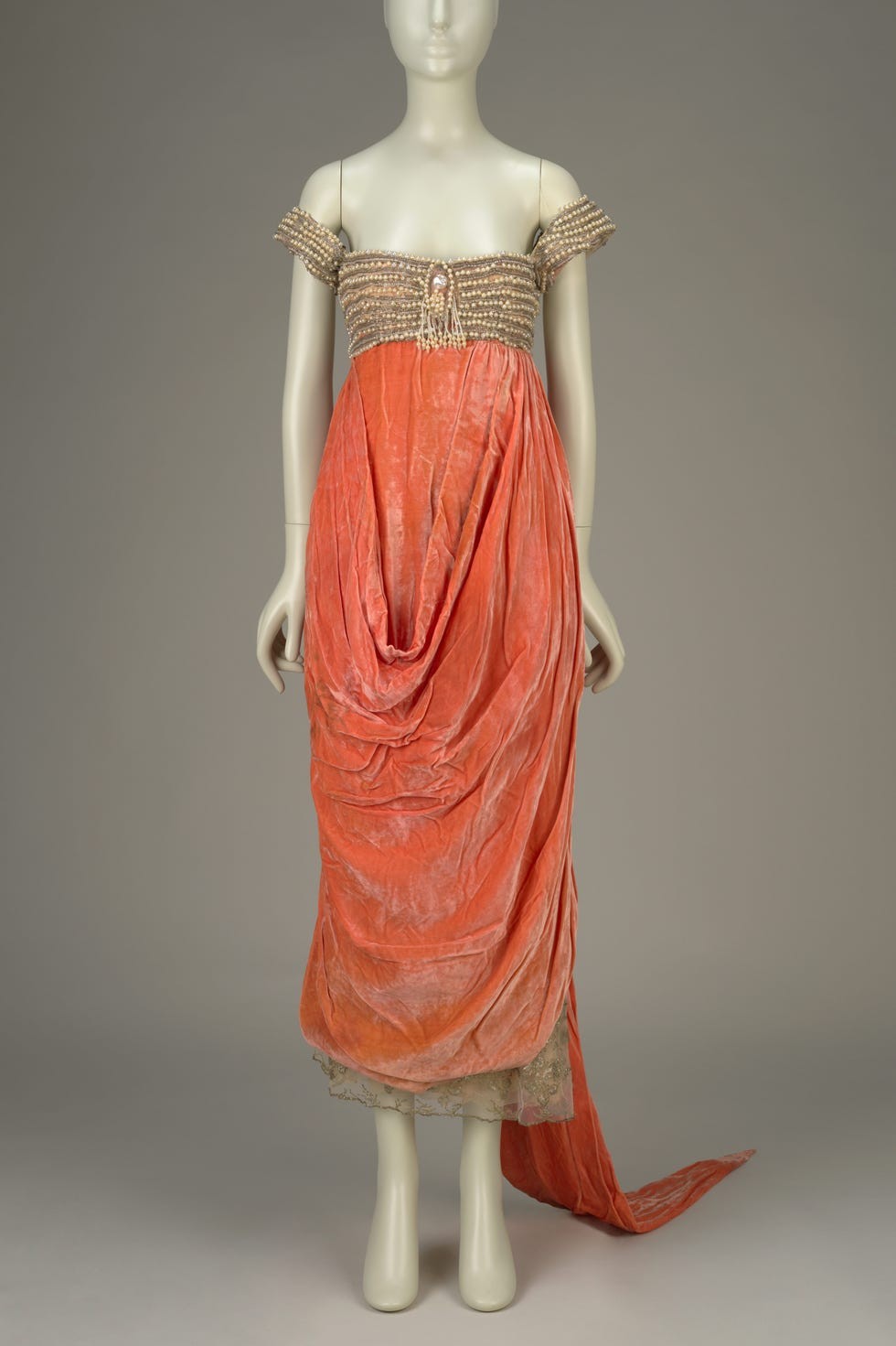 Elegant coral dress with off-the-shoulder sleeves and intricate beading, displayed on a mannequin against a neutral background.
