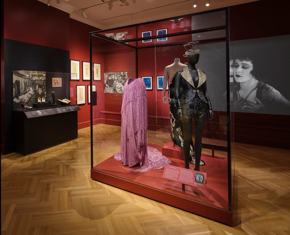 Fashion exhibit featuring vintage costumes, display cases, and framed artwork in a museum setting.