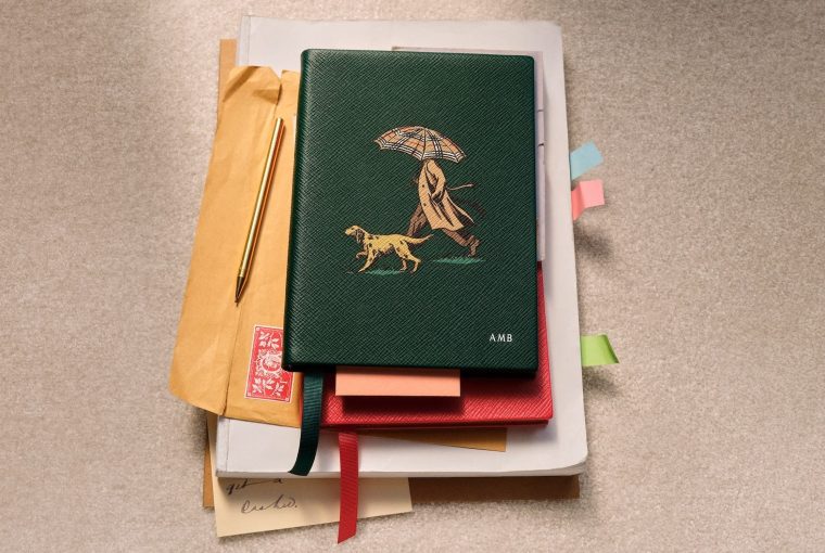 A green notebook with a dog and person under an umbrella, surrounded by papers and a gold pen.
