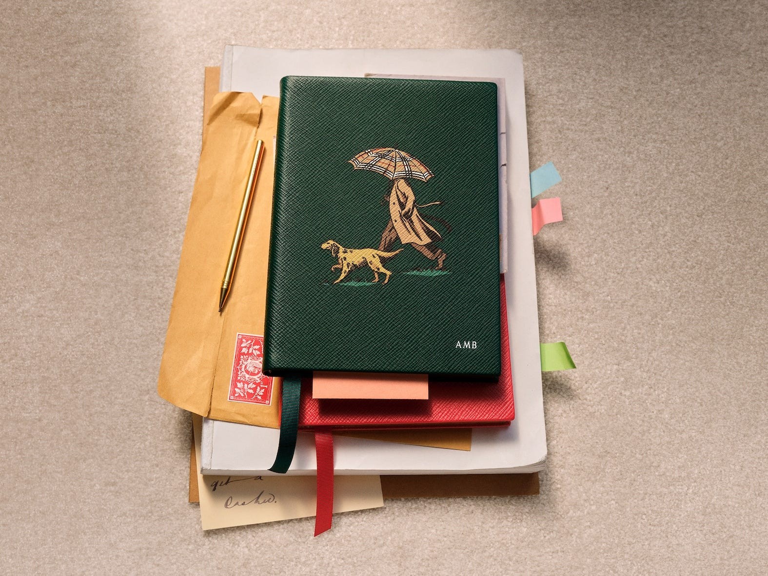 A green notebook with a dog and person under an umbrella, surrounded by papers and a gold pen.