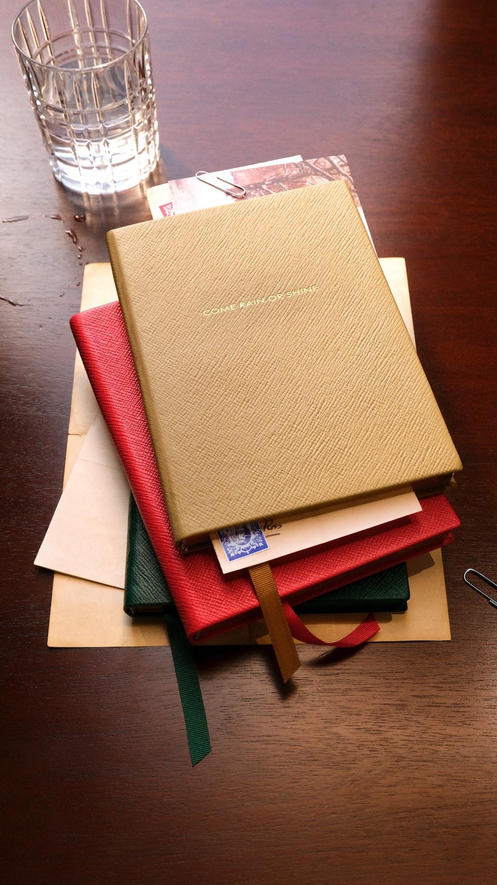 A stack of notebooks with a glass of water and assorted stationery on a wooden table.